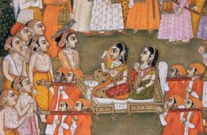 Two richly attired ladies are kneeling on an open palanquin carried by four men in orange uniforms. A crowd of young men stands facing the ladies. All young men and the ladies are wearing long pearl necklaces studded by rubies and pearl earrings. 