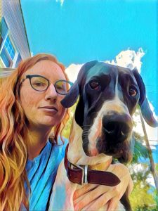 Liz poses with her Great Dane puppy Olive. Liz has long red hair and wears fun blue glasses. Olive looks serious. There is a bright blue sky behind them.