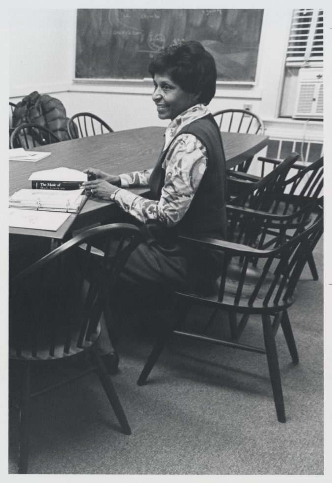 Eileen Southern, smiling, sitting in three quarter profile in a seminar-style classroom, with an open binder of papers and a copy of The Music of Black Americans on the table in front of her.