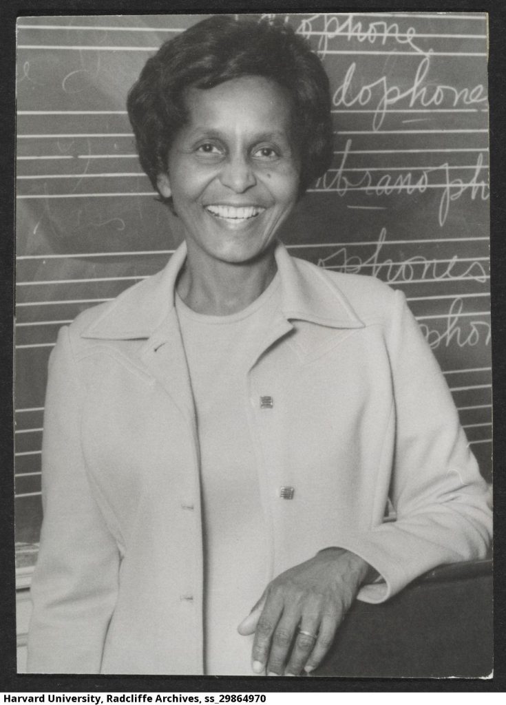Eileen Southern, a Black woman who was a professor at Harvard University, is depicted. Professor Southern is smiling and laughing. She is standing in front of a chalkboard. Professor Southern is wearing a two-piece suit.