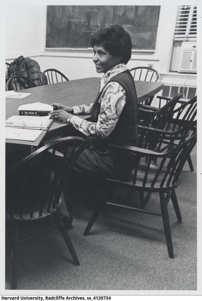 Eileen Southern, a Black woman who was a professor at Harvard University, is depicted. She is sitting at a conference table. Her book The Music of Black Americans rests in front of her. She is wearing a sleeveless dress and a printed blouse. Professor Southern is smiling.